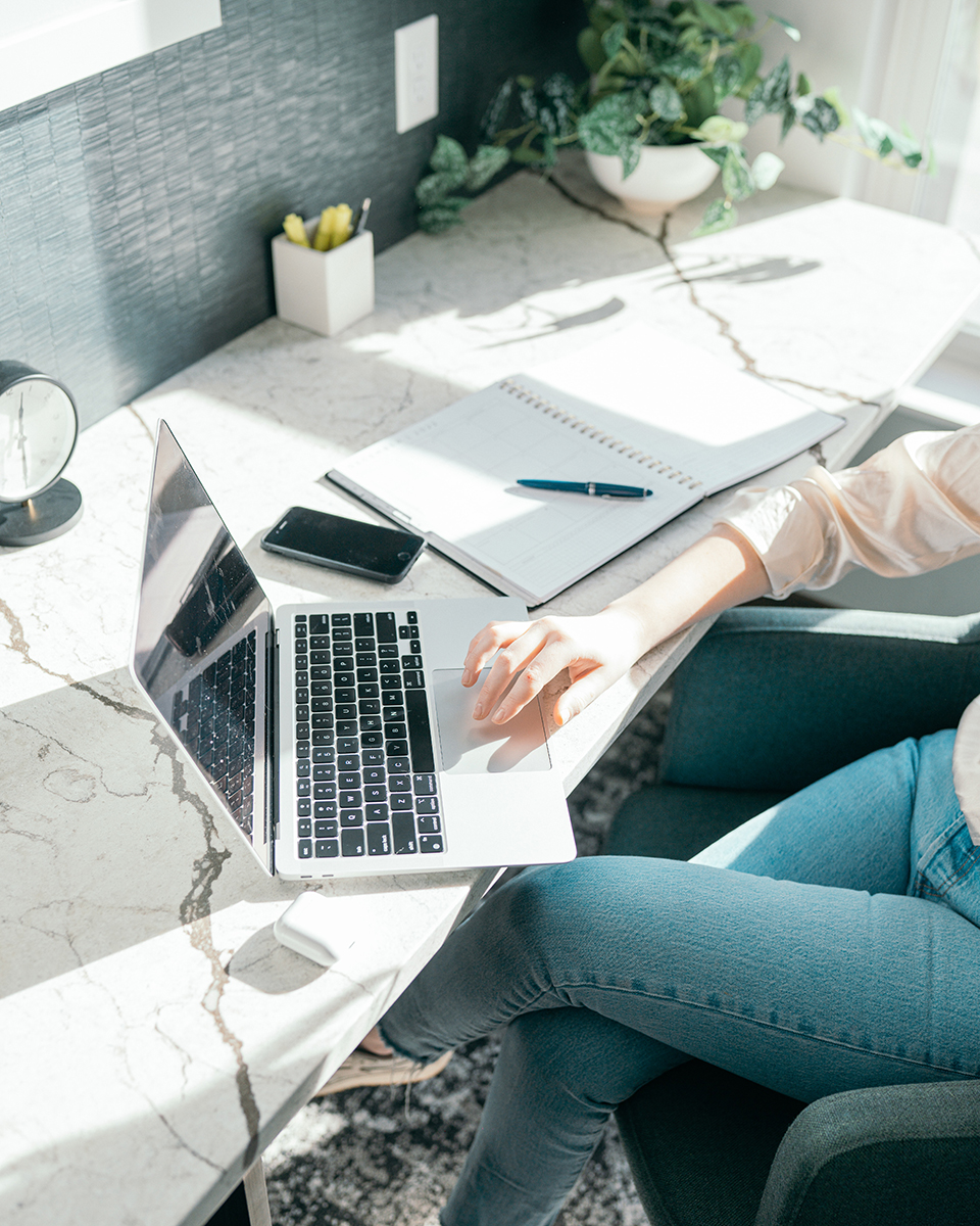 Kvinna sitter vid ett skrivbord med en laptop, en mobil, en almanacka, en penna och en krukväxt.