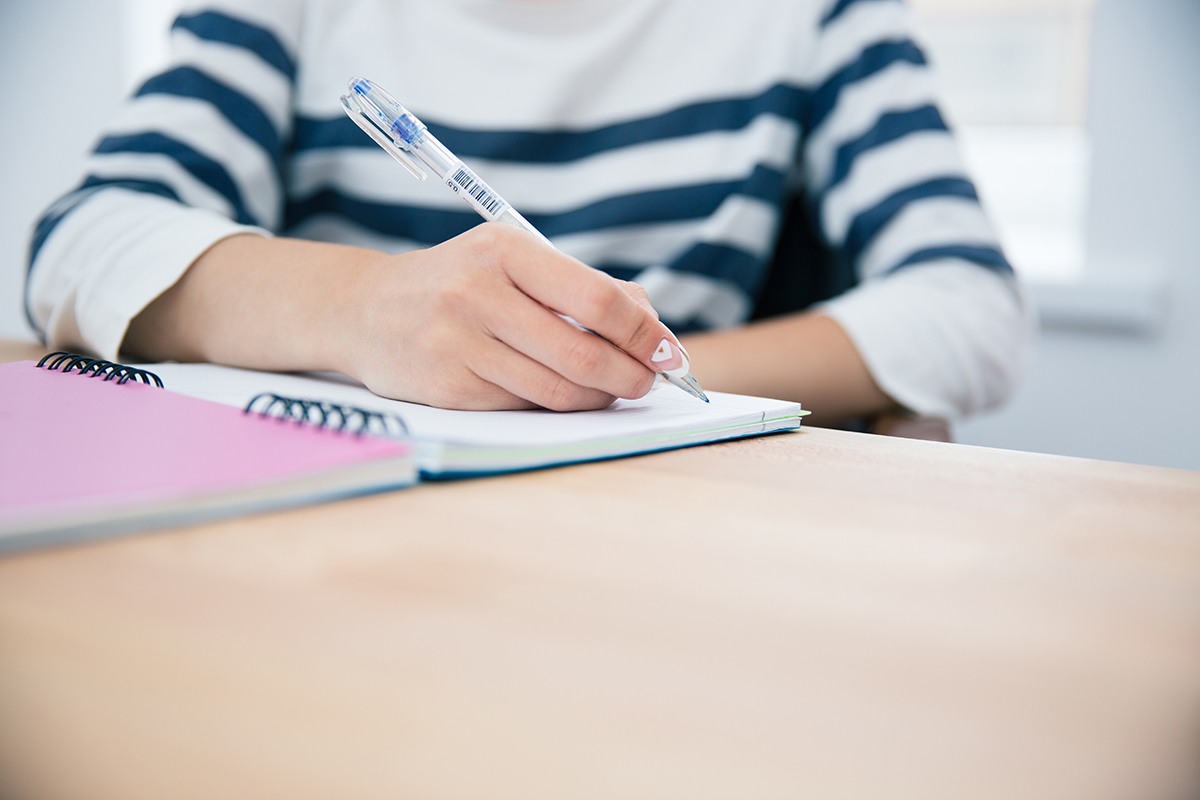 En beskuren bild av en person som sitter vid ett bord och skriver i en anteckningsbok.