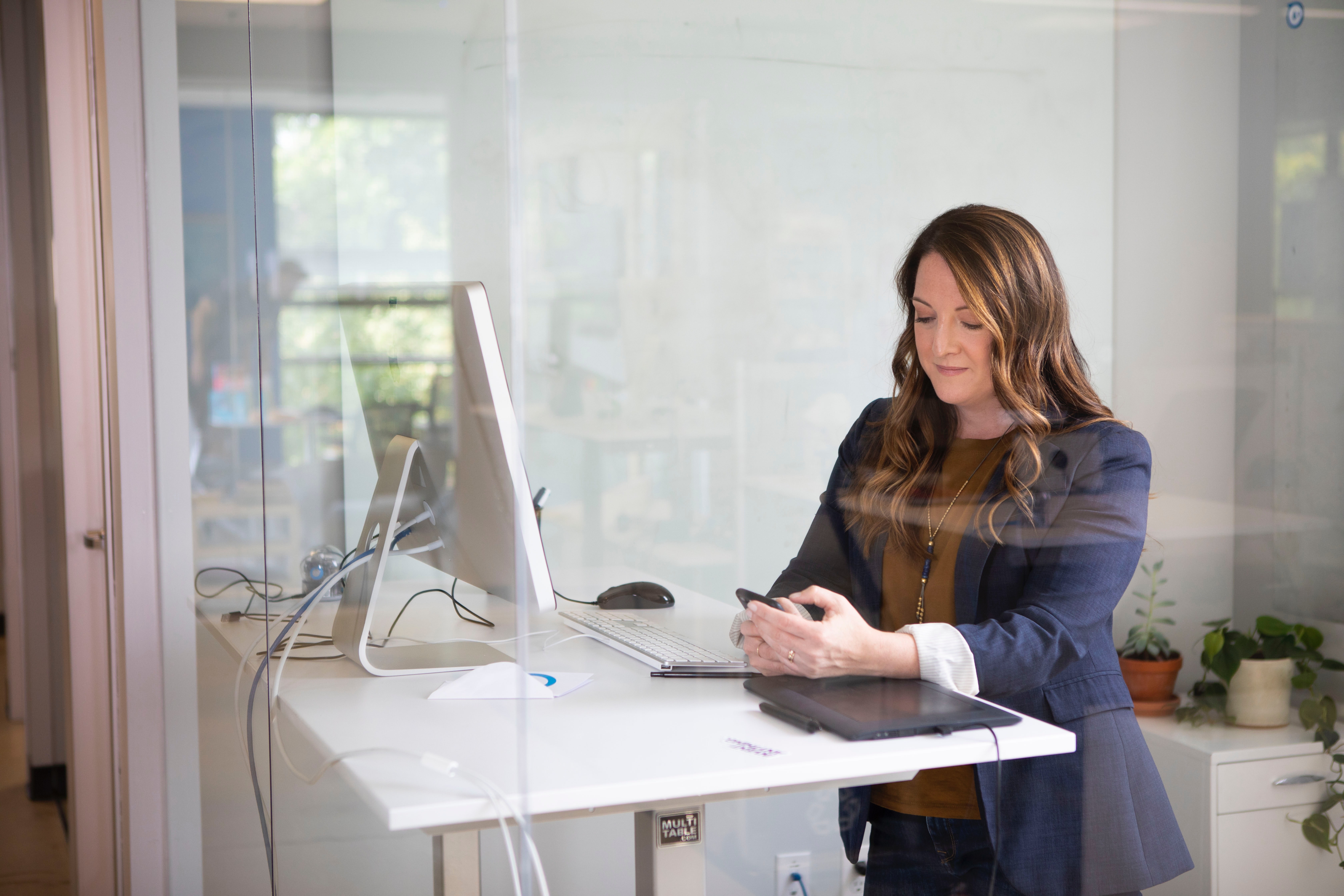 Kvinna står vid ståbord med dator, laptop och penna och tittar på en mobil som hon håller i.
