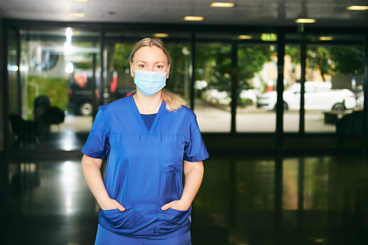 Kvinnlig fysioterapeut iklädd munskydd och bussarong  står i sjukhusentré och tittar rakt in i kameran.